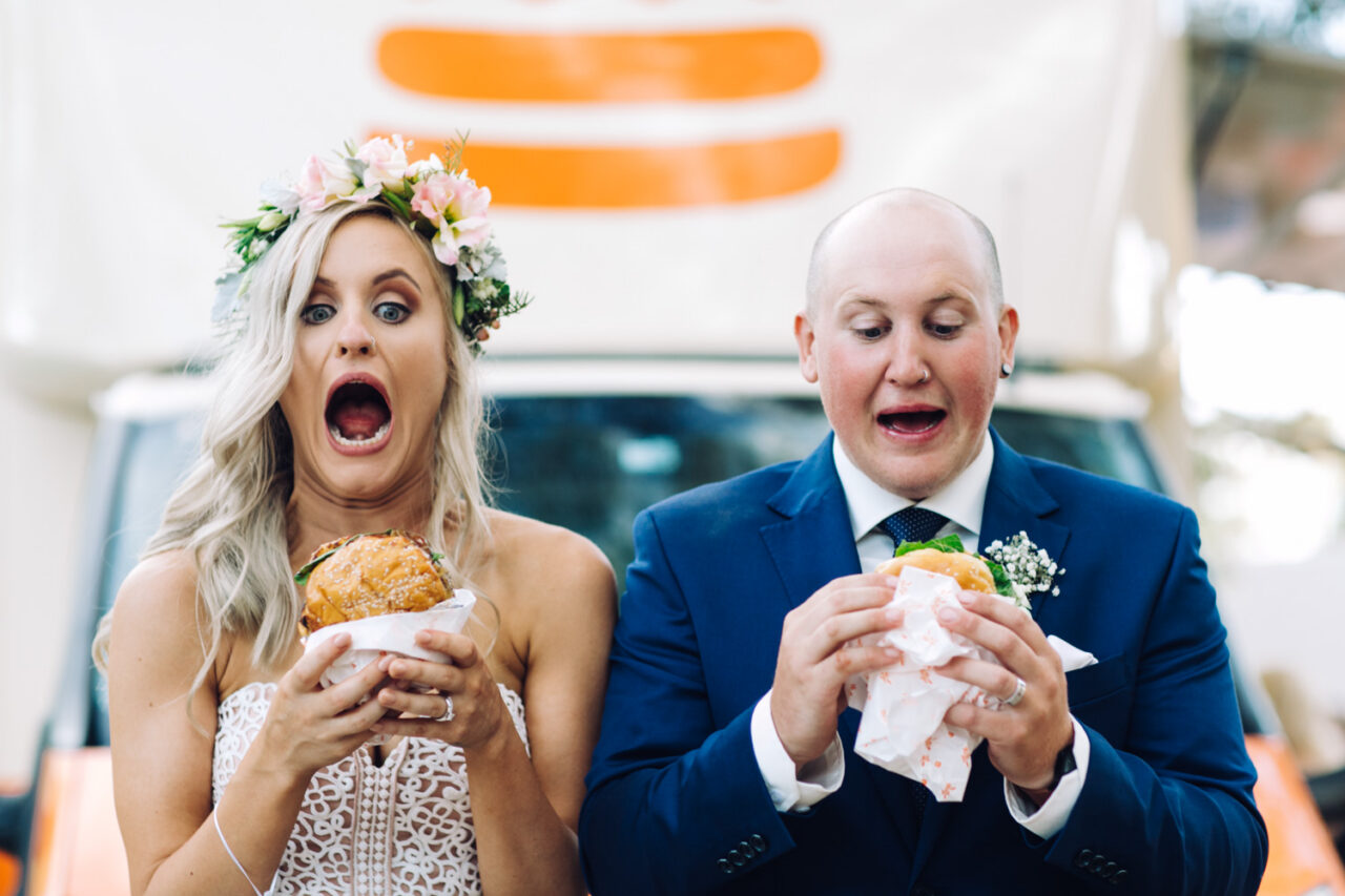 Couple reacting with surprise while eating burgers on their wedding day
