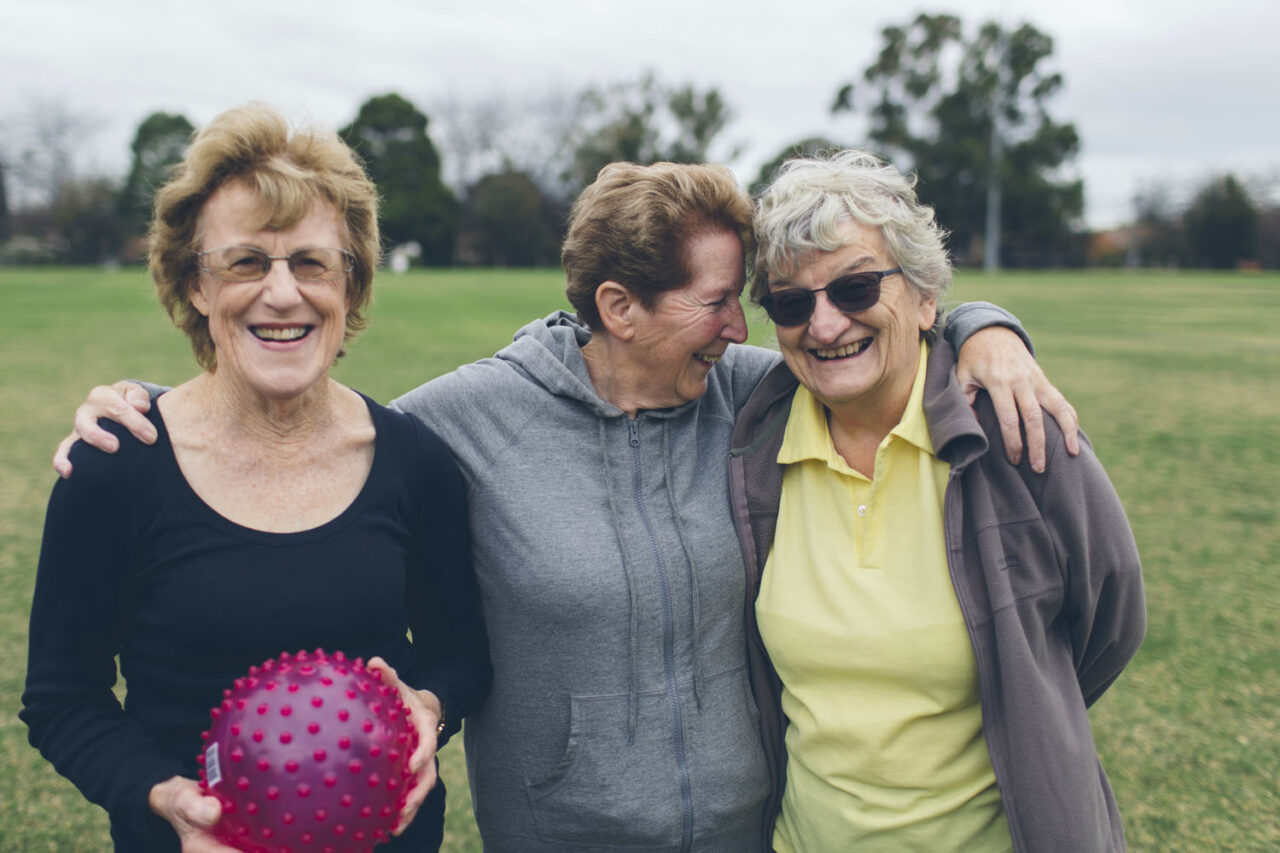 Three older women smiling and embracing after a YMCA fitness session outdoors