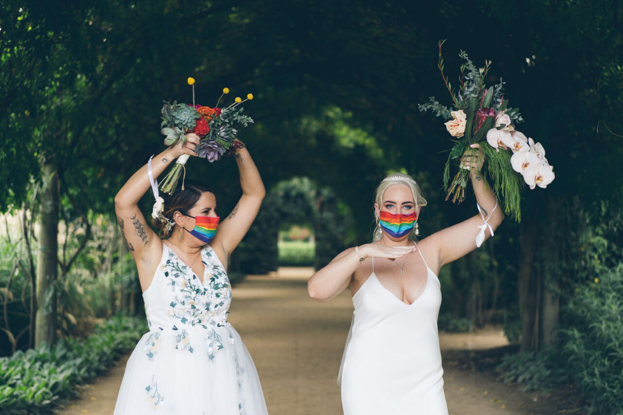 Two brides celebrating with bouquets and rainbow masks beneath the garden archway at Alowyn Gardens in the Yarra Valley.