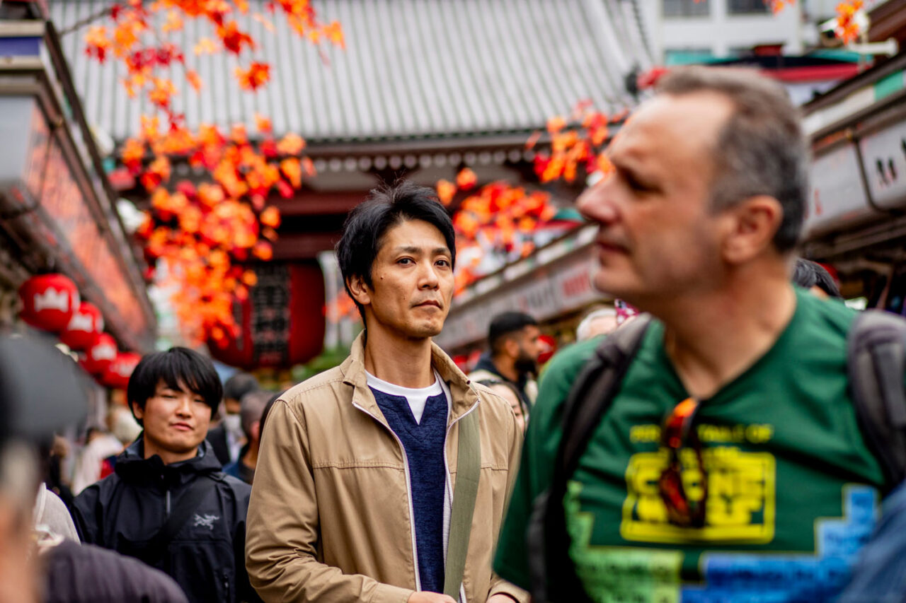 Man walking through a crowded Asakusa street framed by red autumn leaves near Sensoji Temple