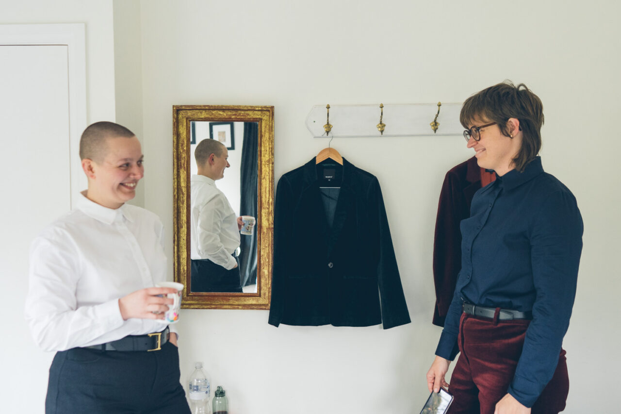 Naomi and Bec helping each other get dressed in tailored suits before their Daylesford wedding.