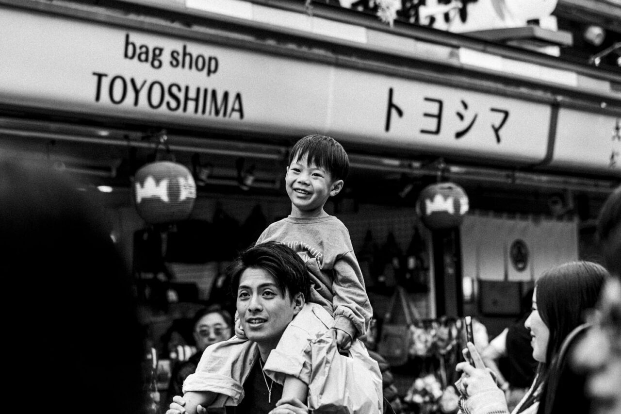 Child sitting on his father’s shoulders smiling in a busy Asakusa street scene in black and white