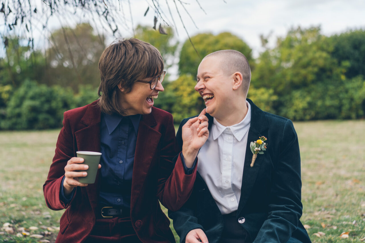 Naomi and Bec laughing together outdoors while holding coffee cups on their wedding day in Daylesford.