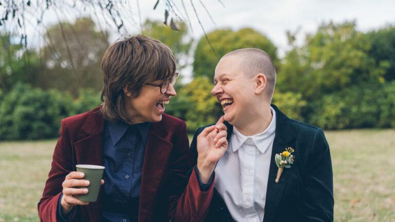 Naomi and Bec laughing together outdoors while holding coffee cups on their wedding day in Daylesford.