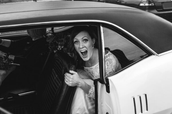 Bride laughing excitedly as she steps out of a vintage car on her wedding day, captured in a candid documentary moment.