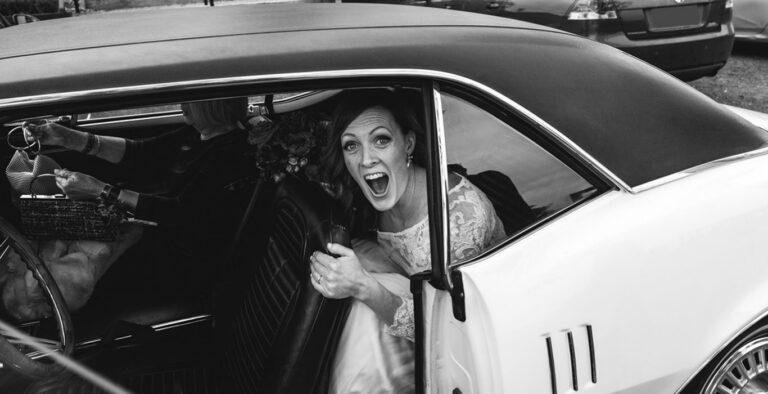 Bride laughing excitedly as she steps out of a vintage car on her wedding day, captured in a candid documentary moment.
