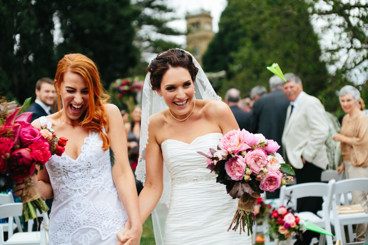 Two brides walk back down the aisle laughing and holding hands after their wedding ceremony.