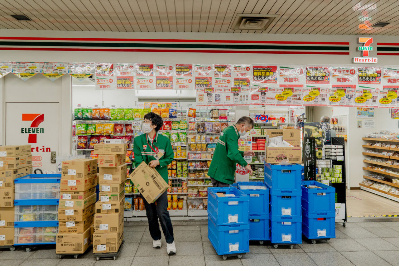 wo 7-Eleven staff members in green uniforms stocking shelves outside a busy Japanese convenience store
