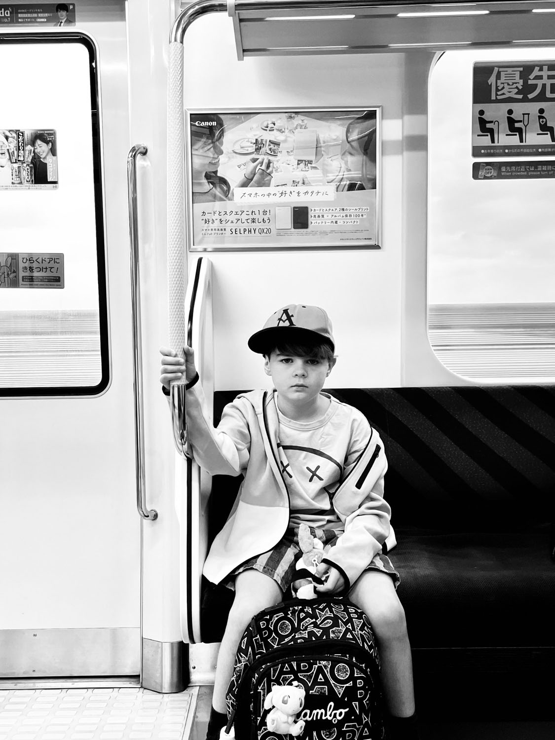 Young boy sitting alone on a Tokyo train carriage holding the rail, backpack at his feet, black and white documentary photograph