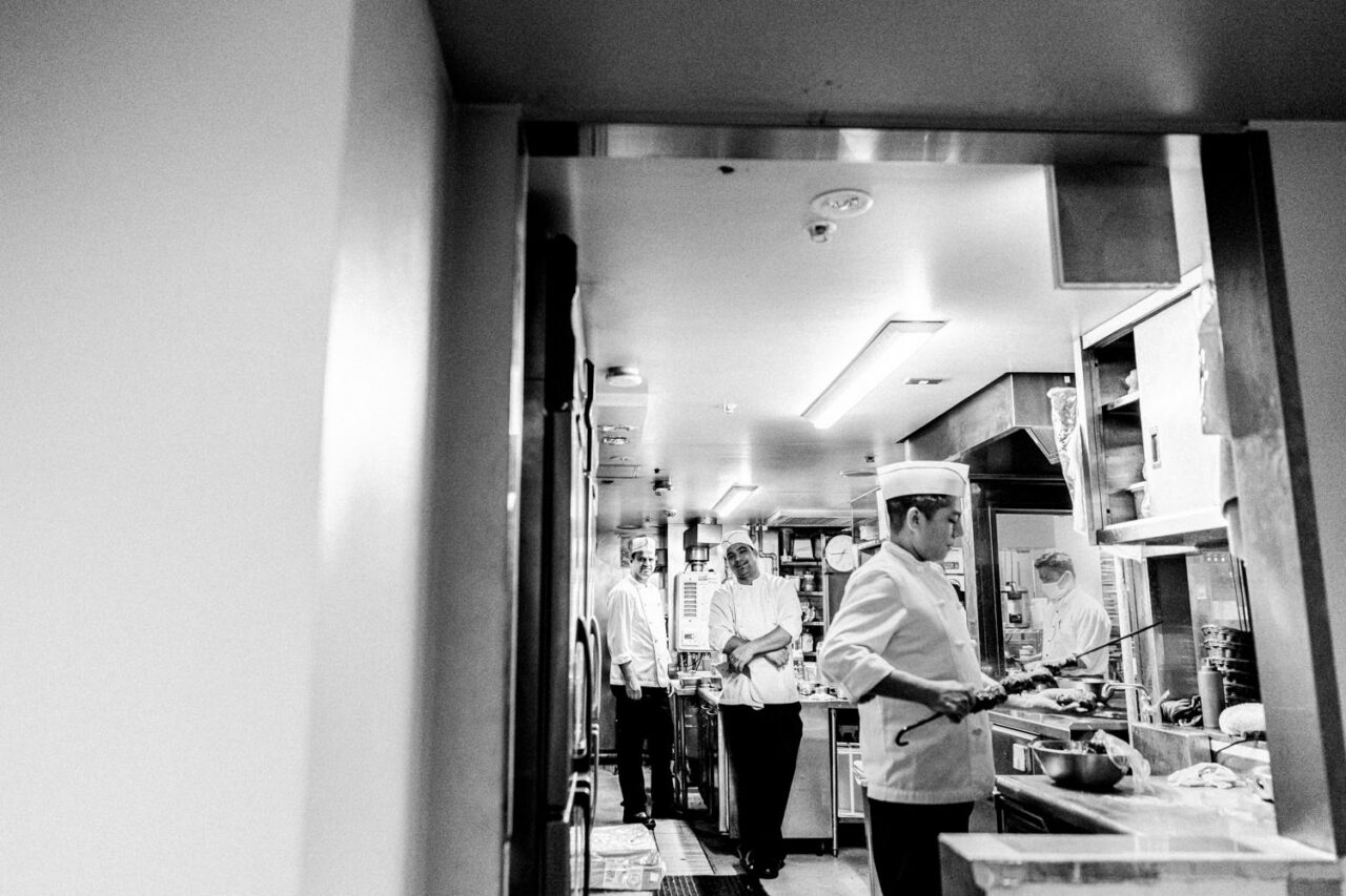 Documentary black and white photograph of chefs working and watching inside a Japanese restaurant kitchen, viewed through a doorway
