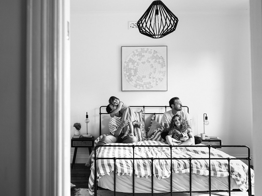 A family of four on their bed, photographed through a doorway in black and white, capturing a quiet and real moment at home during a Your Story session, Surf Coast Victoria 2025.