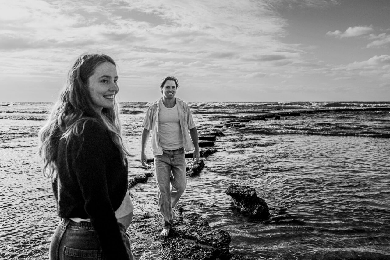Black and white image of pregnant woman smiling back over her shoulder as her partner walks toward her across the rock shelf, ocean behind them