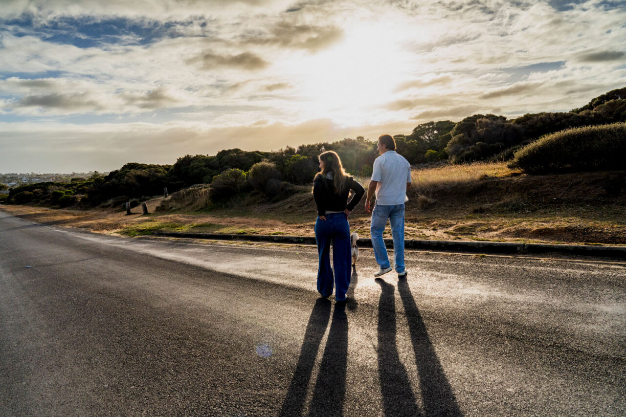 Pregnant couple and their dog walking toward the beach on the Surf Coast at sunrise, long shadows cast behind them on the road