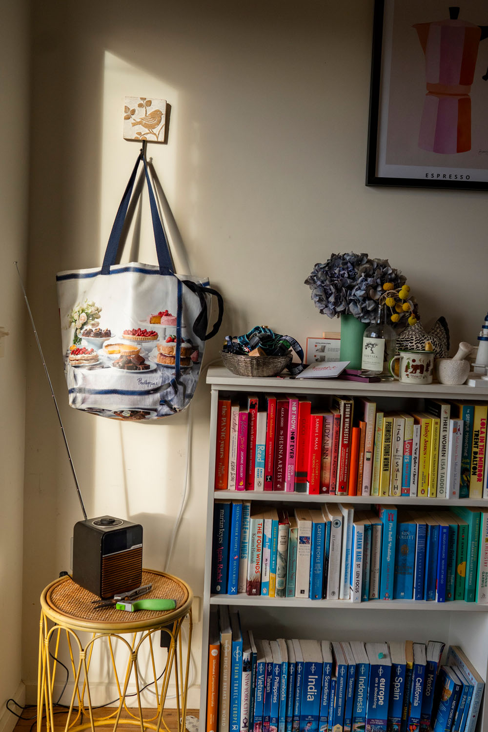 Corner of a living room with a colourful bookshelf of travel books, espresso poster print above, tote bag on a bird hook, small radio on a rattan stool