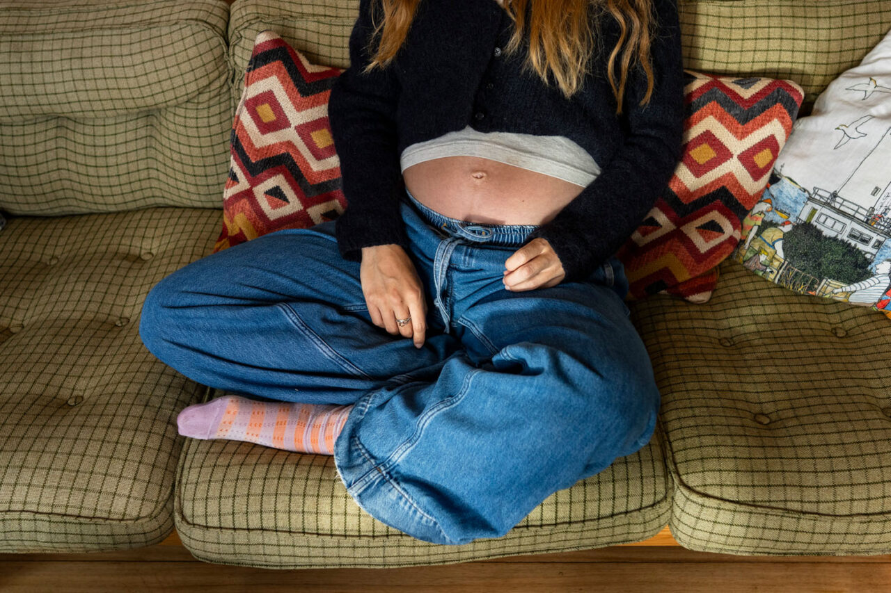 Pregnant woman sitting cross legged on the couch at 29 weeks, belly showing, wearing jeans and a black cardigan with pink printed socks