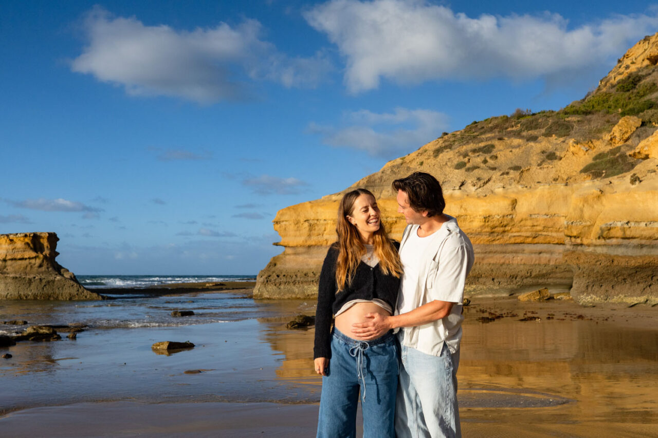 Man holding pregnant woman's belly while she smiles with her eyes closed, sandstone cliffs and blue sky on the Surf Coast behind them