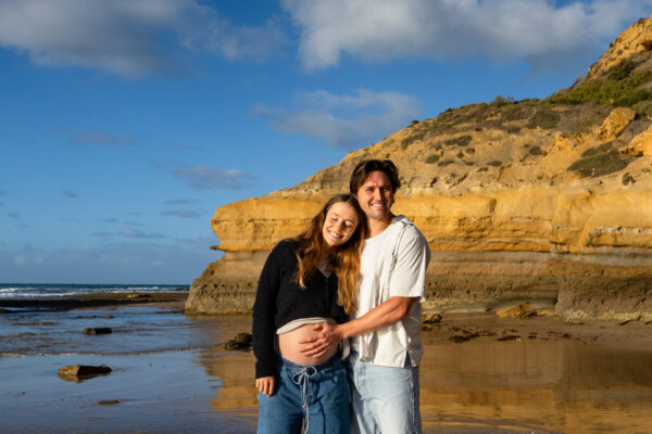 Pregnant couple smiling together on the Surf Coast beach at low tide, sandstone cliffs glowing amber behind them, man's hand resting on her 29-week belly
