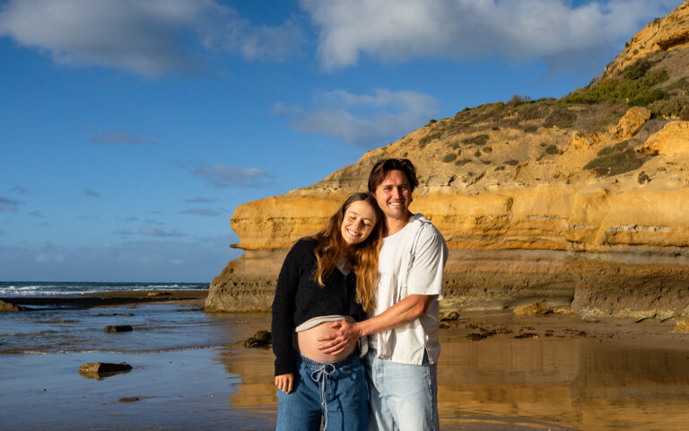 Pregnant couple smiling together on the Surf Coast beach at low tide, sandstone cliffs glowing amber behind them, man's hand resting on her 29-week belly