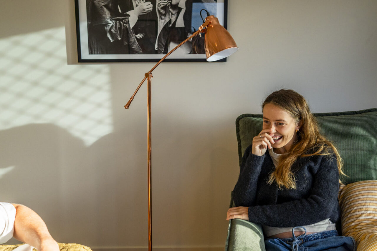Pregnant woman laughing in a green armchair with a copper floor lamp behind her, morning light casting diamond shadows on the wall, framed black and white print above
