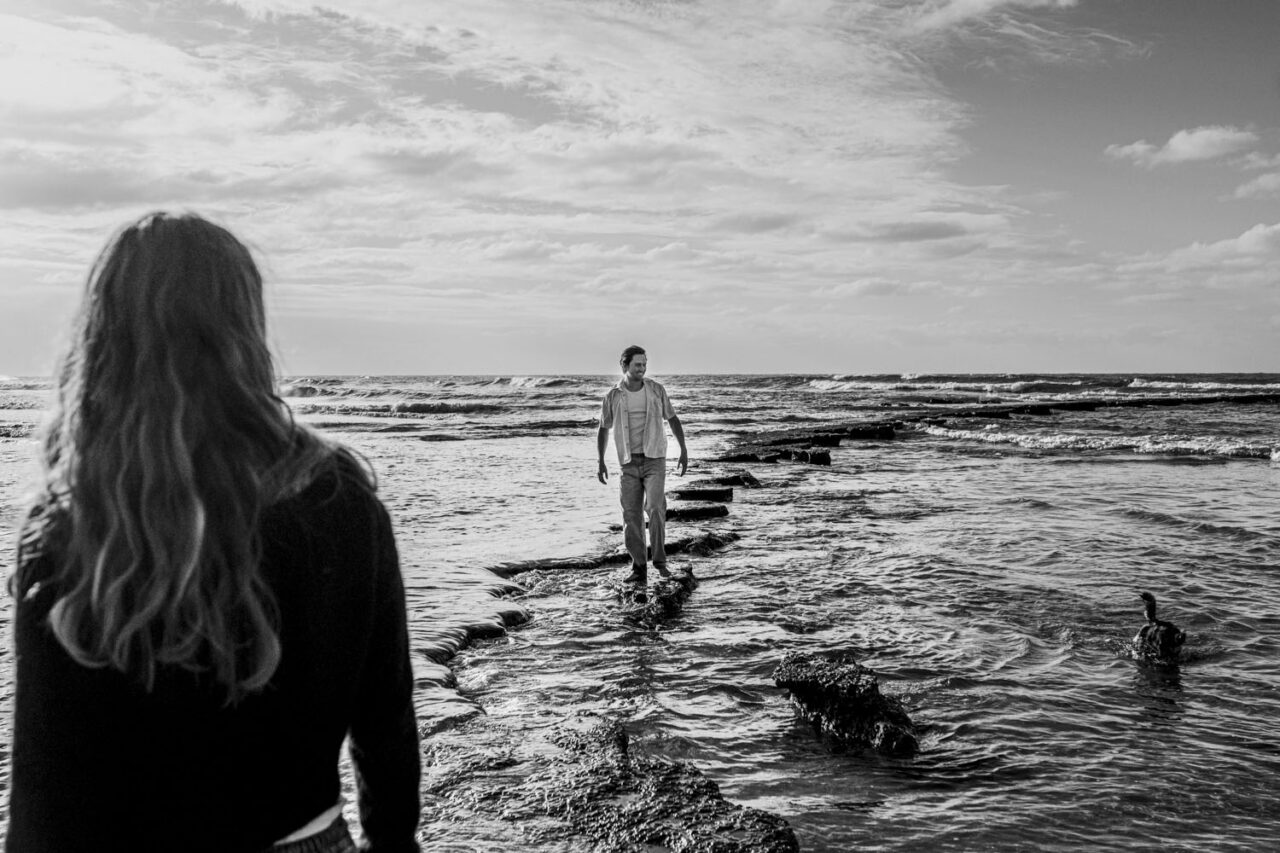 Black and white image of a man walking across the rock shelf toward the camera on the Surf Coast, his partner's hair and shoulders in soft focus in the foreground, dog visible in the water to the right, wide ocean and sky behind