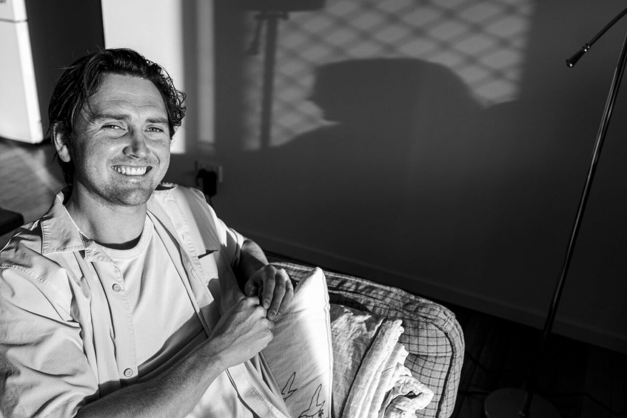 Black and white portrait of a man smiling relaxed on a couch in morning light, bird print cushion beside him, lamp and shadow behind