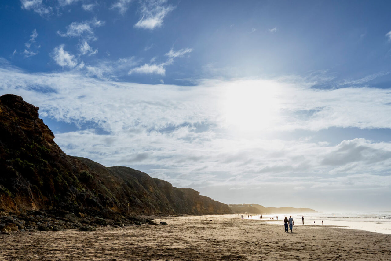 Wide landscape shot of the Surf Coast beach at low tide with sandstone cliffs on the left and couple small in the frame walking toward the light
