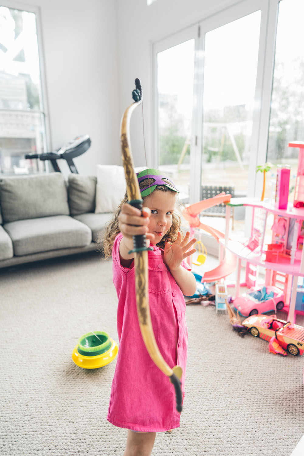 A young girl in a pink dress and ninja mask aims a toy bow and arrow at the camera with fierce concentration, surrounded by toys, captured during a documentary family photography session in Geelong 2025.
