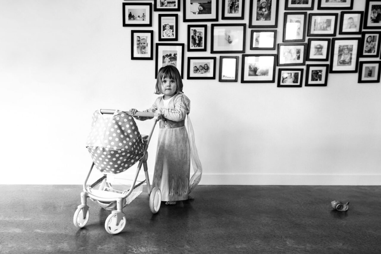 A young girl in an Elsa costume pushes a polka dot doll pram across a concrete floor in front of a large gallery wall of family photos, captured in black and white during a documentary family photography session in Geelong 2025.