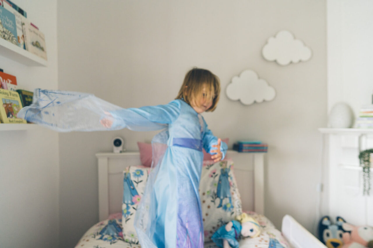 A young girl in an Elsa costume spins on her bed, arms outstretched and dress blurring with movement, in a soft light bedroom during a documentary family photography session in Geelong 2025