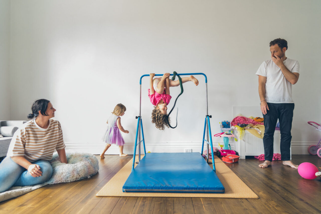 A young girl hangs upside down on an indoor gymnastics bar while her mother watches with delight and her father looks on with mild concern, captured during a documentary family photography session in Geelong 2025.