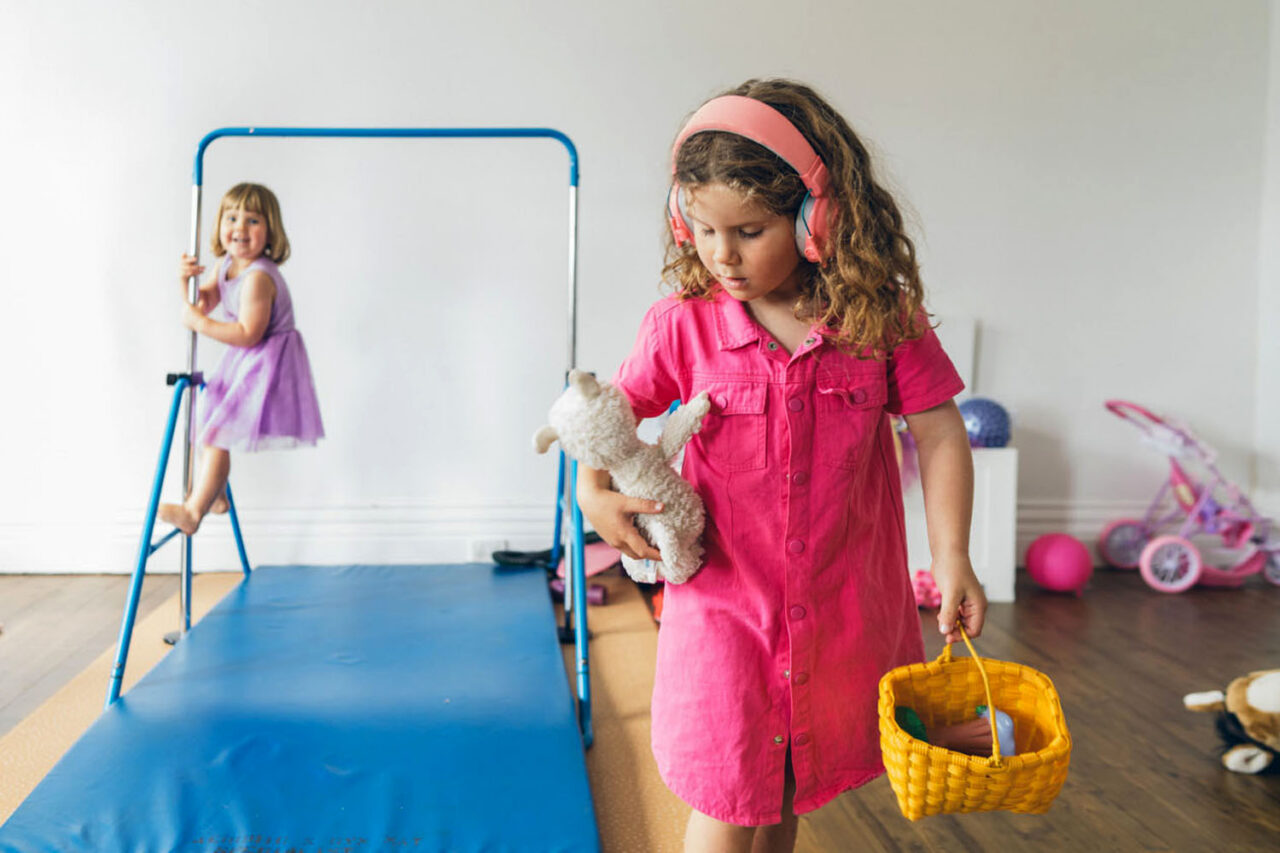 A girl in pink with headphones and a yellow basket walks past the gymnastics bar where her younger sister perches, while their father watches from the background, captured during a documentary family photography session in Geelong 2025.