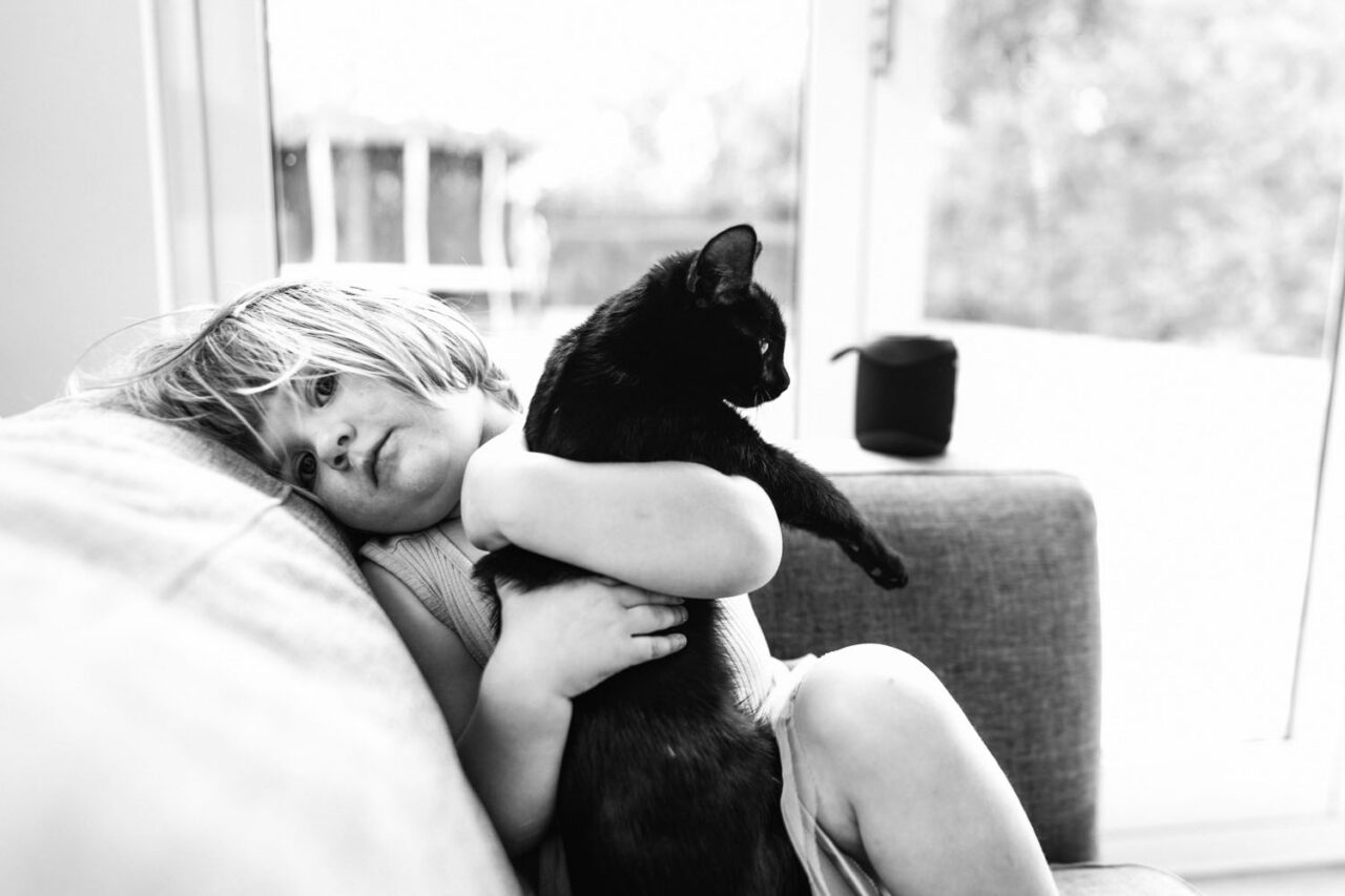 A young girl lies on the couch in black and white, arms wrapped around the family black cat who gazes out the window, captured during a documentary family photography session in Geelong 2025.