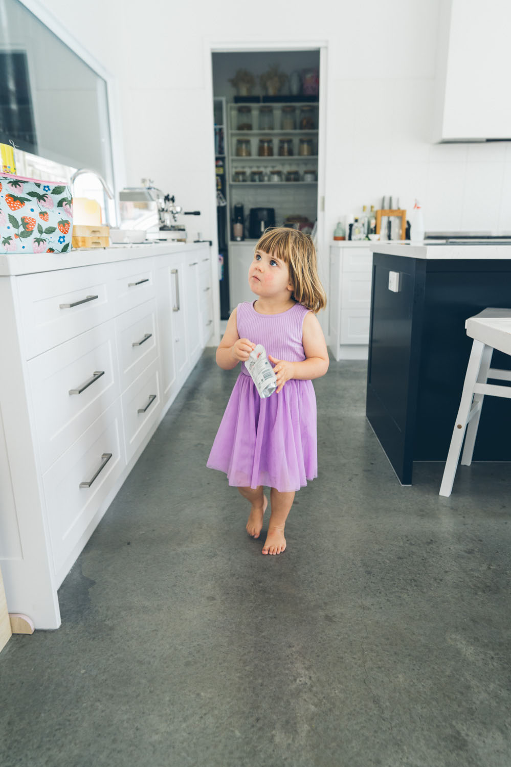 A young girl in a purple tulle dress stands barefoot in a modern white kitchen, looking upward with a curious expression, captured during a documentary family photography session in Geelong 2025.