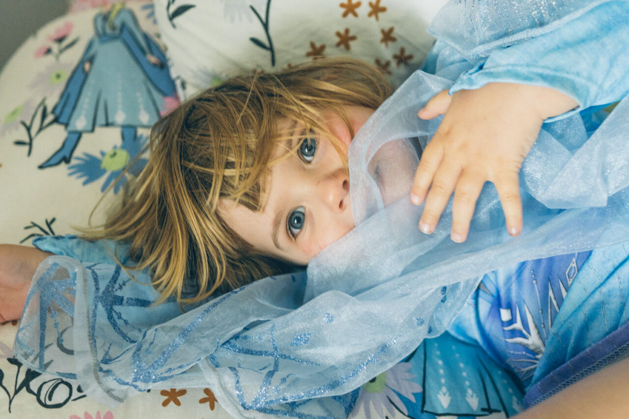 A young girl lies on her back on a floral bed wearing an Elsa costume, looking directly into the camera with wide blue eyes and messy hair, captured during a documentary family photography session in Geelong 2025.