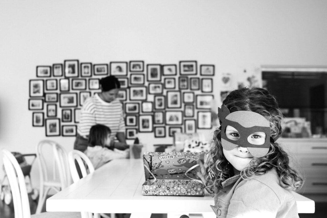 A young girl wearing a superhero mask sits at a dining table looking sideways at the camera with a knowing smile, her mother and sister visible in the background in front of a large gallery wall, captured in black and white during a documentary family photography session in Geelong 2025.