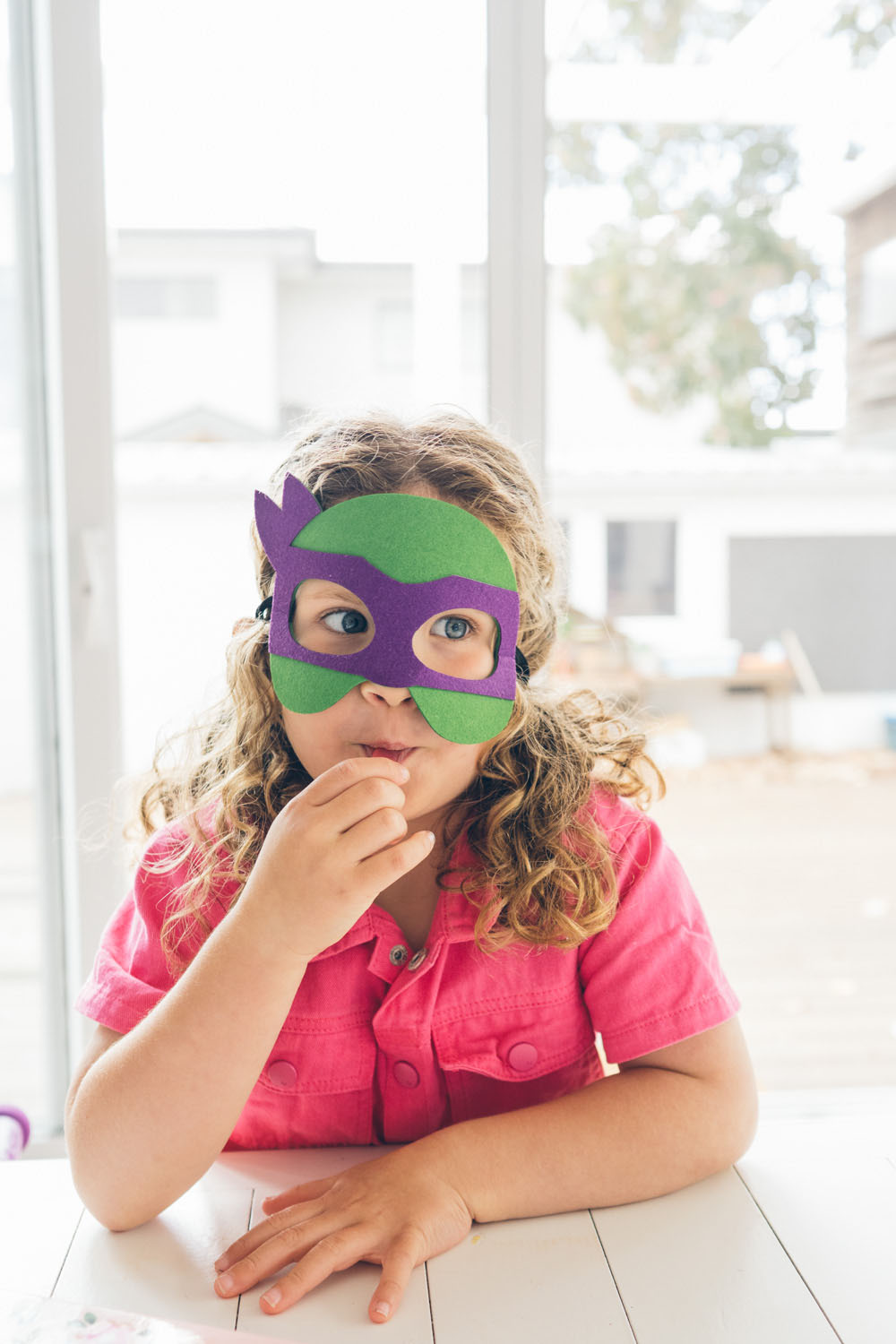 A young girl with curly hair wearing a green and purple ninja mask rests her chin on her arms at a white dining table, eyeing the camera with quiet amusement, captured during a documentary family photography session in Geelong 2025.