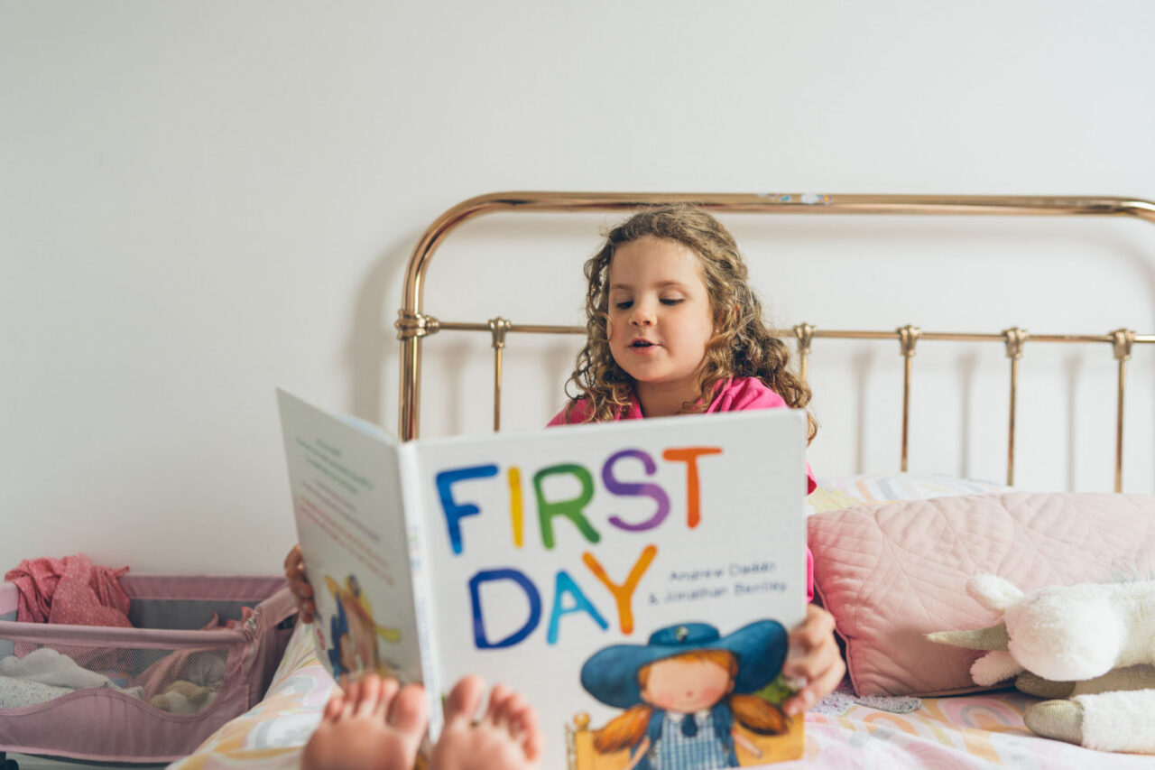 A young girl with curly hair sits on a brass bed reading a picture book aloud, captured during a documentary family photography session in Geelong 2025.