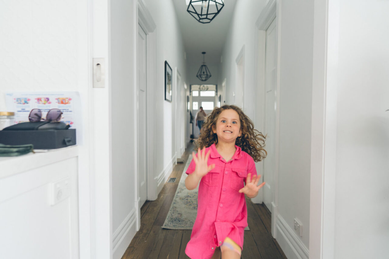 A young girl with curly hair tears down a long bright hallway with her hands outstretched and a wild grin on her face, captured during a documentary family photography session in Geelong 2025.