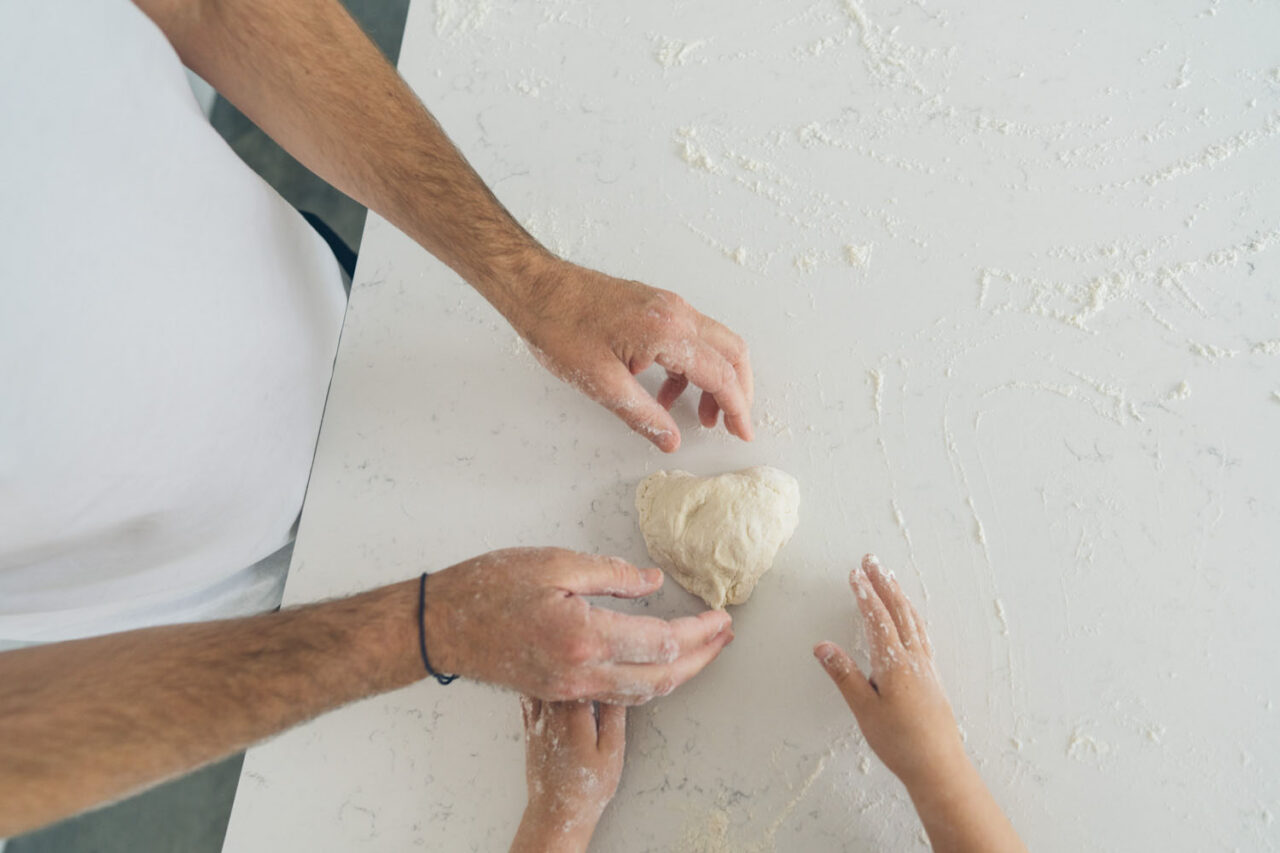 Overhead shot of a father's hands shaping dough beside a small child's hand reaching in to help, on a flour-covered kitchen bench, captured during a documentary family photography session in Geelong 2025.