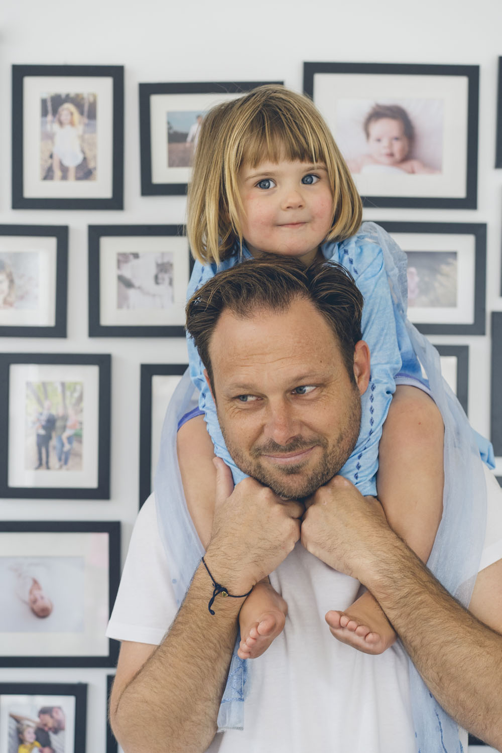 A young girl sits on her father's shoulders in front of a large gallery wall of family portraits, both looking at the camera with quiet confidence, captured during a documentary family photography session in Geelong 2025.