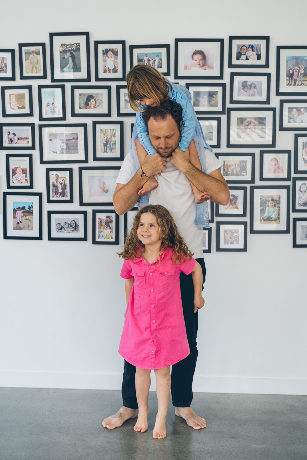 A father stands in front of a gallery wall with his youngest daughter draped across his shoulders and his eldest standing proudly in front, all three looking at the camera, captured during a documentary family photography session in Geelong 2025.