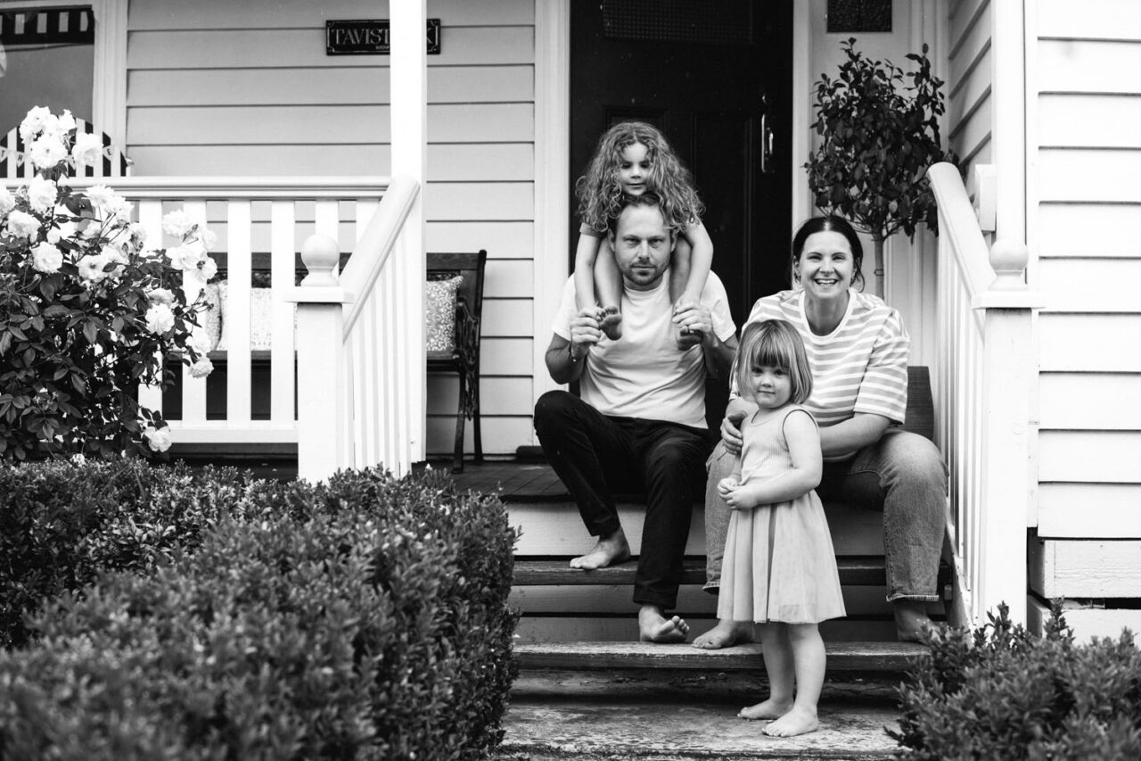 A family of four sits on the front steps of a heritage home in black and white, one daughter on her father's shoulders, the other leaning against her mother, captured during a documentary family photography session in Geelong 2025.