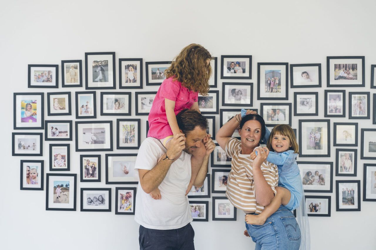 A family of four laughs in front of a large gallery wall of framed family photos, kids on parents shoulders, captured during a documentary family photography session in Geelong 2025.