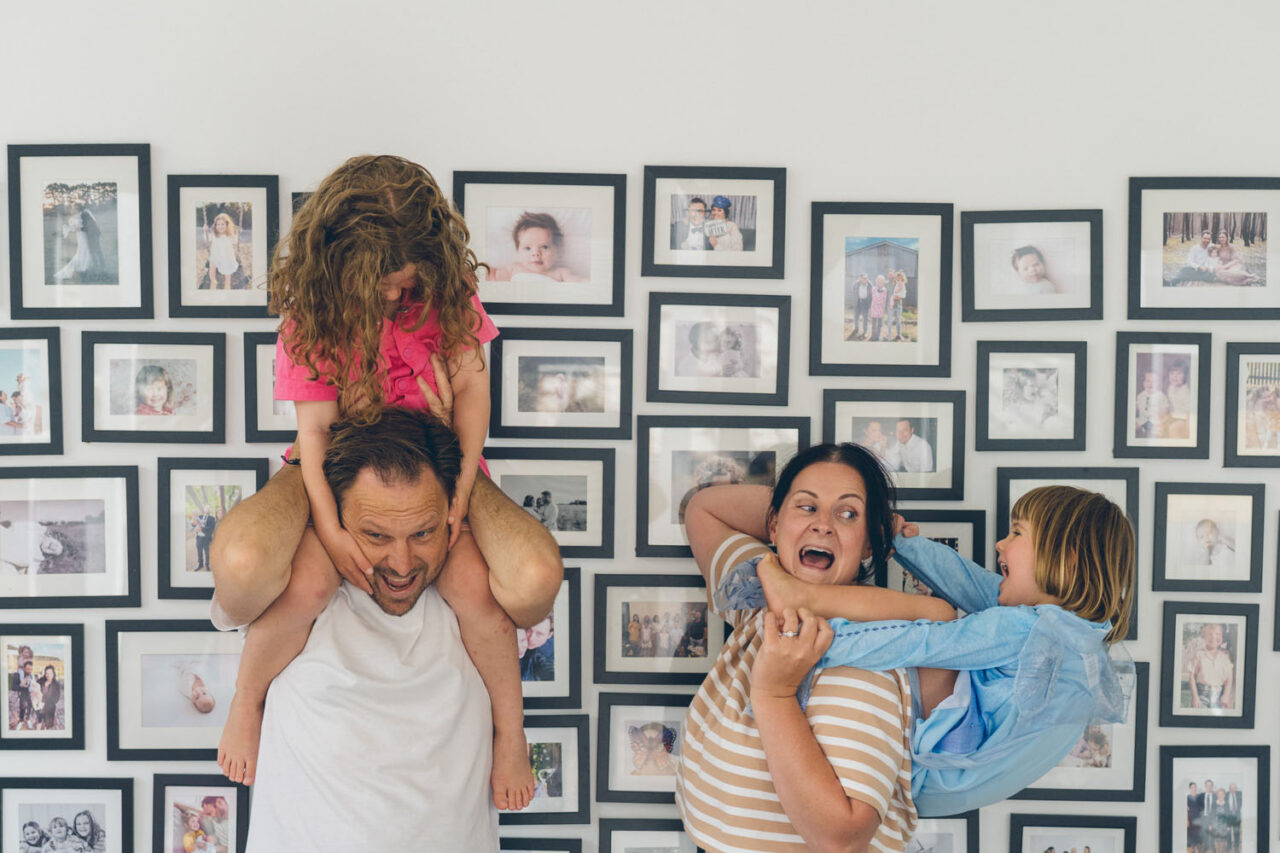 A mother laughs with her mouth wide open as her daughter clings to her back and her partner grins while their other daughter hangs off his shoulders, captured in front of a gallery wall during a documentary family photography session in Geelong 2025.