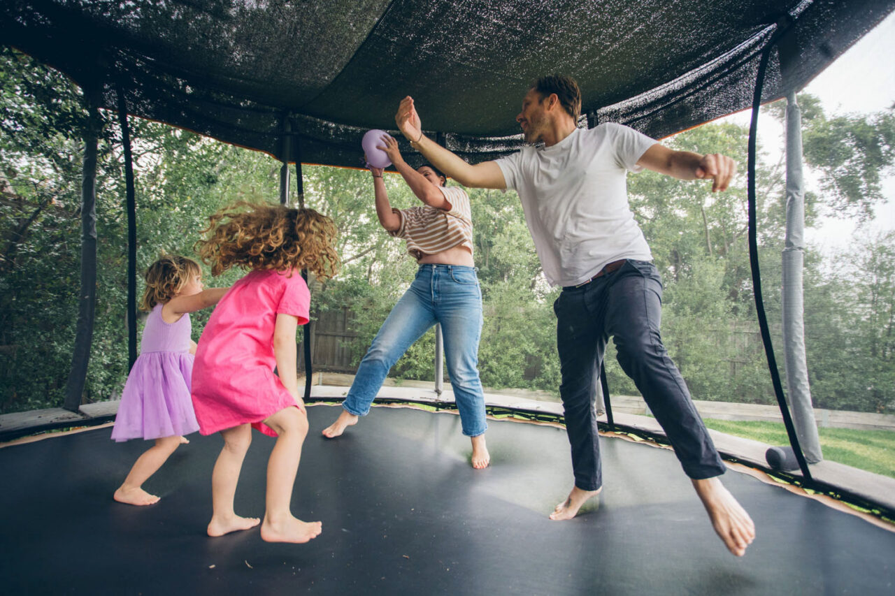 A family of four bounces together on a backyard trampoline, arms out and mid-air, captured during a documentary family photography session in Geelong 2025.
