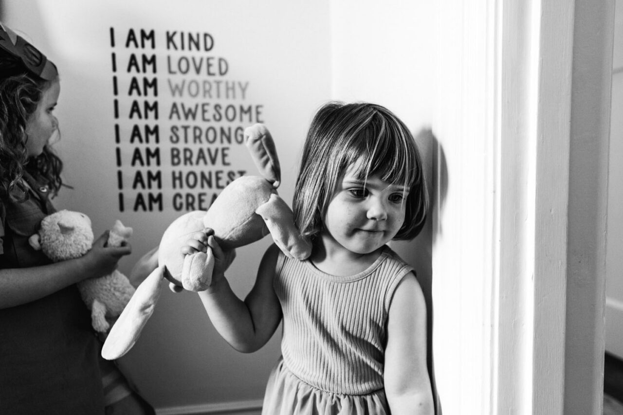 A young girl leans against a wall printed with the words I Am Kind, I Am Loved, I Am Worthy, holding a stuffed bunny, while her sister stands beside her, captured in black and white during a documentary family photography session in Geelong 2025.