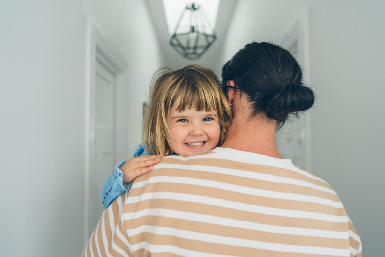 A mother carries her young daughter down a bright hallway, the child grinning over her shoulder at the camera, captured during a documentary family photography session in Geelong 2025.