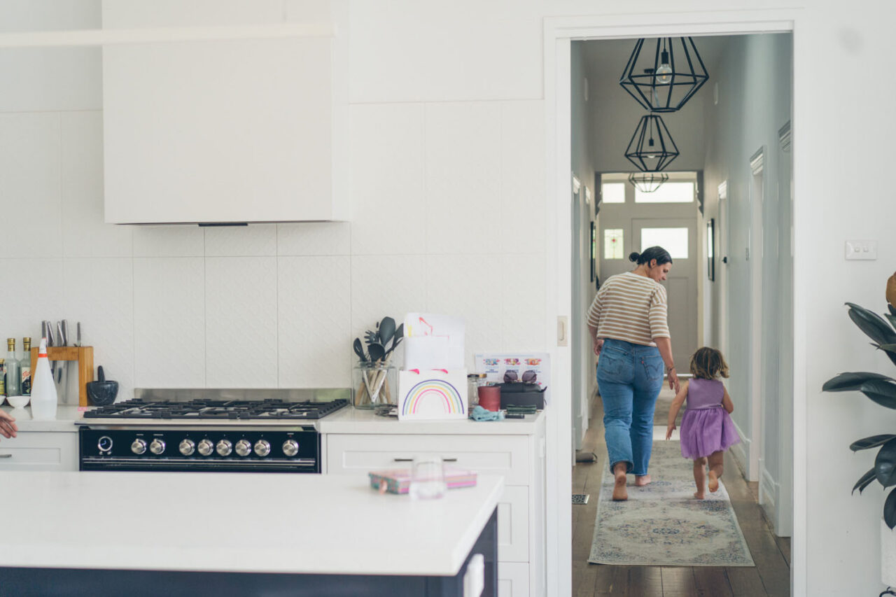A mother and young daughter walk hand in hand from the kitchen into a bright hallway, shot from across the room during a documentary family photography session in Geelong 2025.