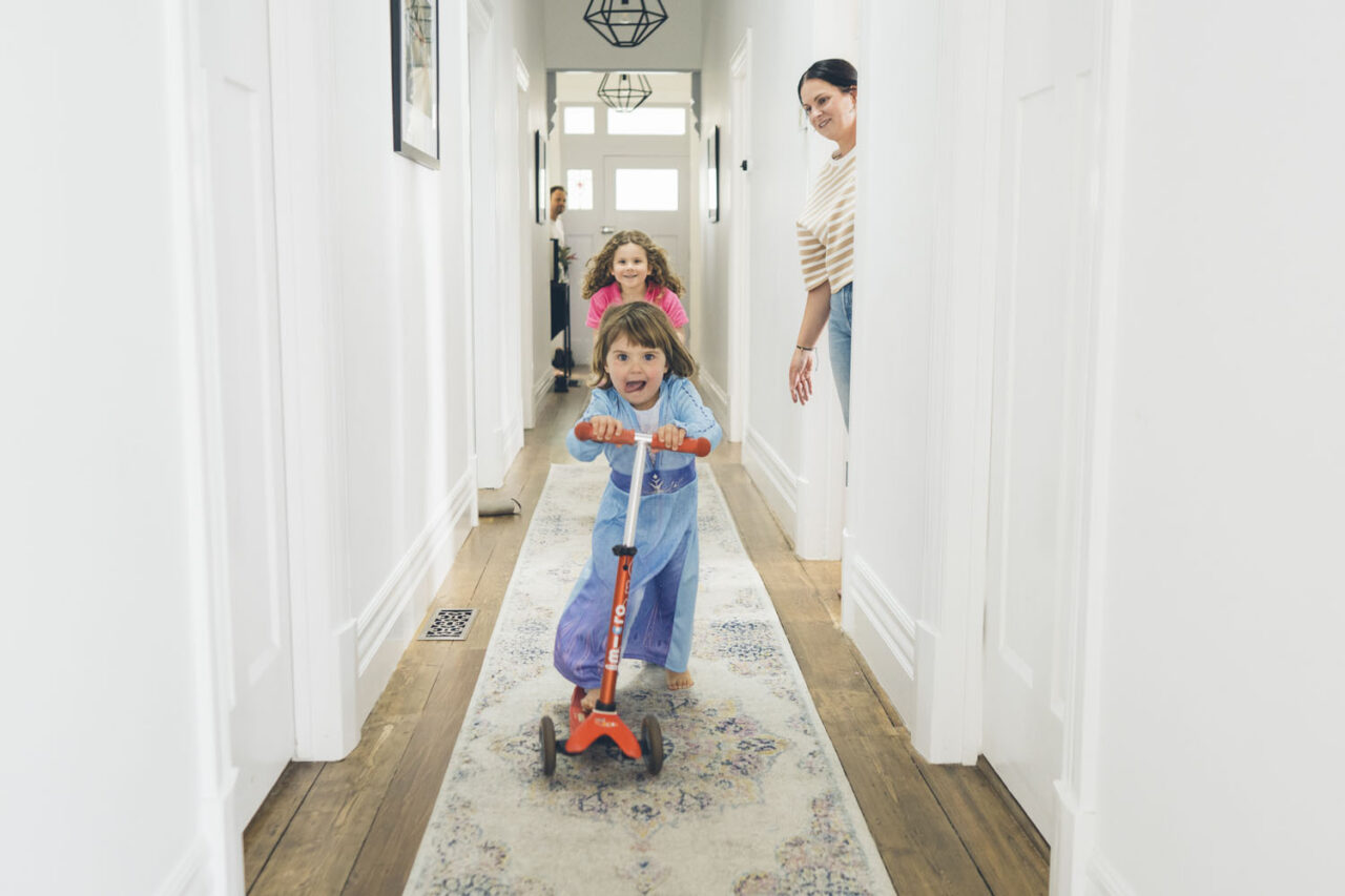 A young girl rides a scooter at full speed down a bright hallway while her sister chases and her mother watches on, captured during a documentary family photography session in Geelong 2025