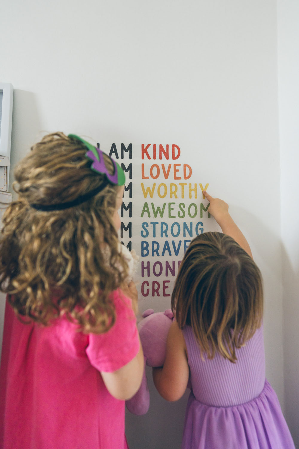 Two sisters stand side by side with their backs to the camera, pointing at a colourful I Am affirmation wall together, captured during a documentary family photography session in Geelong 2025.